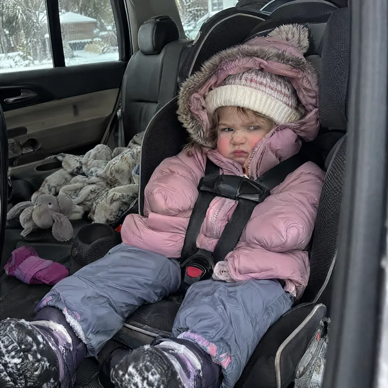 photo of a bundled-up toddler, visibly uncomfortable, squeezed into a car seat while wearing a thick winter coat